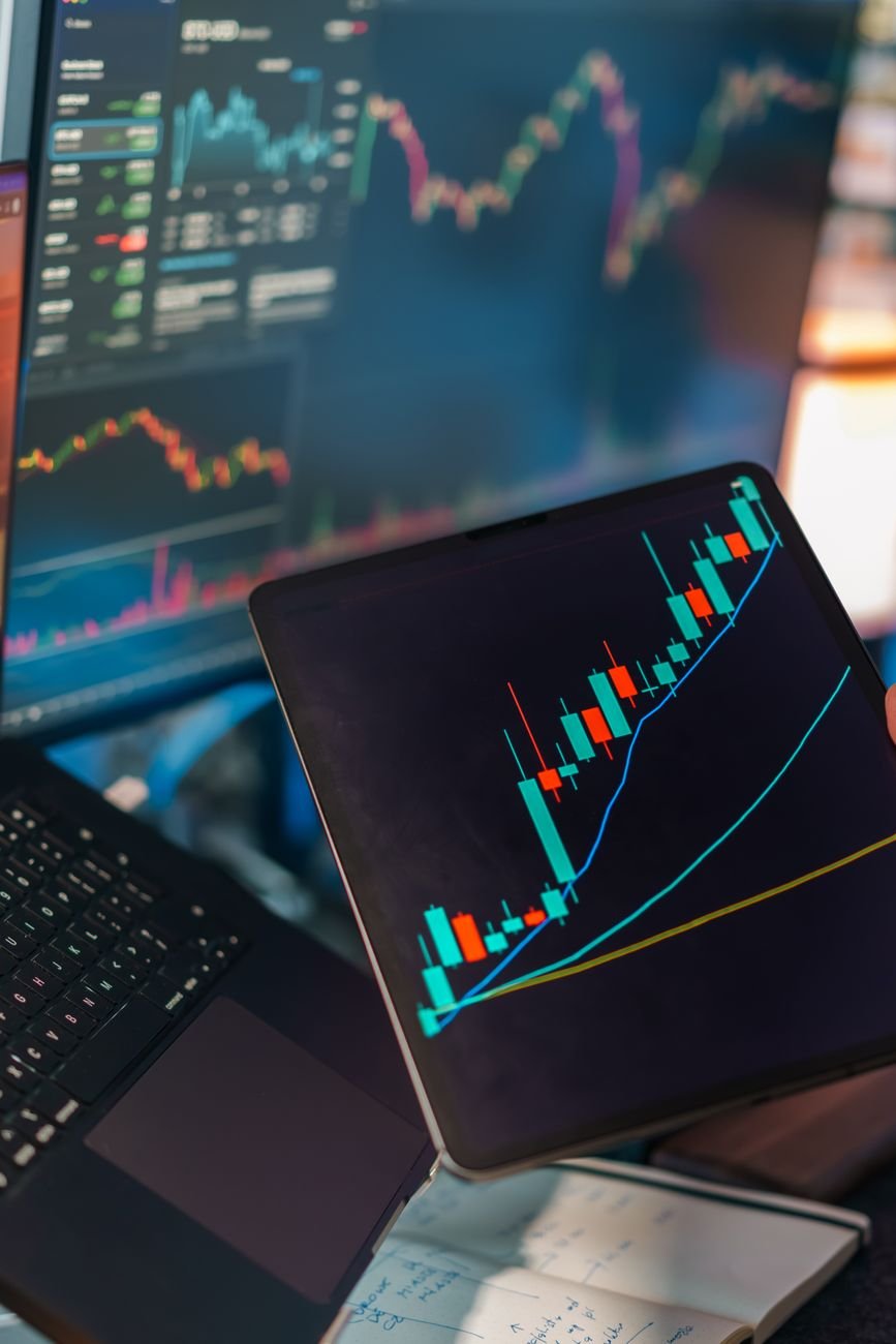 A tablet displaying a candlestick chart with a laptop screen showing stock market data.
